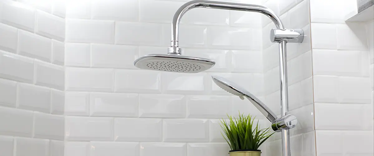 Modern shower with white glossy subway tiles, chrome rain showerhead, handheld shower, and a small green plant on a corner shelf.