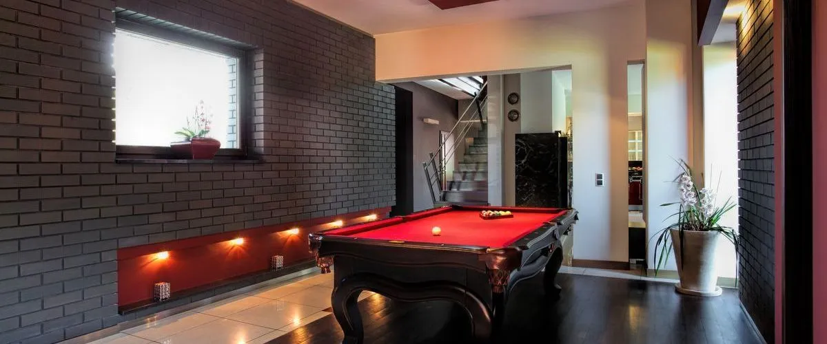 A remodeled basement featuring a stylish game room with a red felt pool table, dark brick accent wall, modern lighting, and contemporary decor, creating a sleek and inviting atmosphere