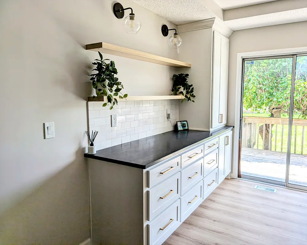 White kitchen with built-in cabinets, floating wooden shelves, black countertop, subway tile backsplash, and sliding glass door.